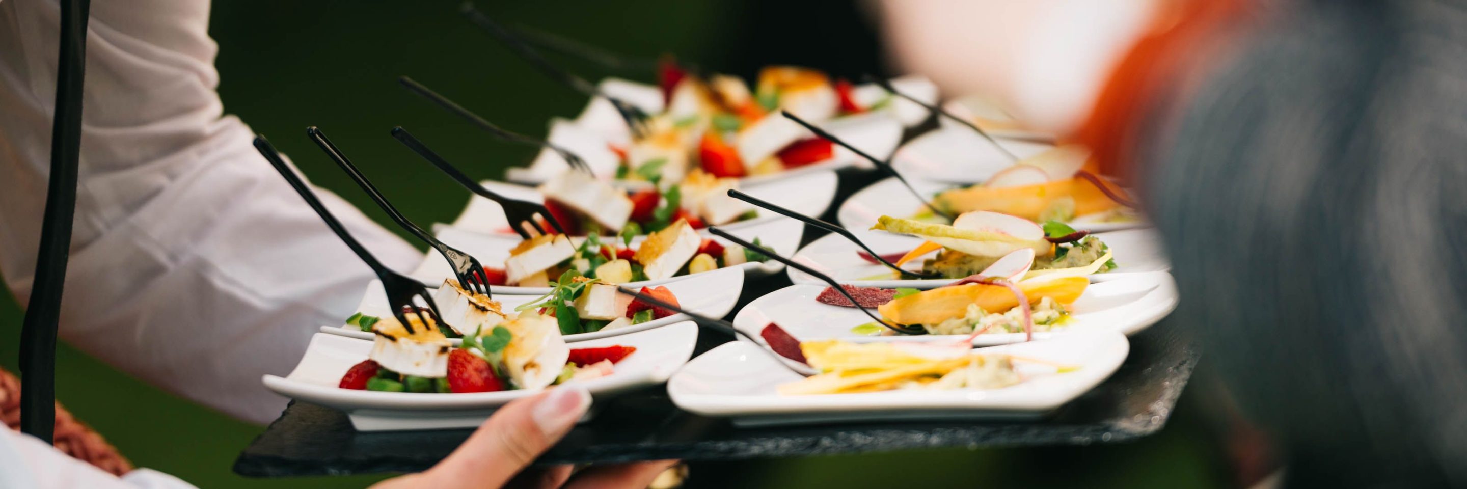 A server carrying a tray of canapés at an outdoor event, guests reaching in